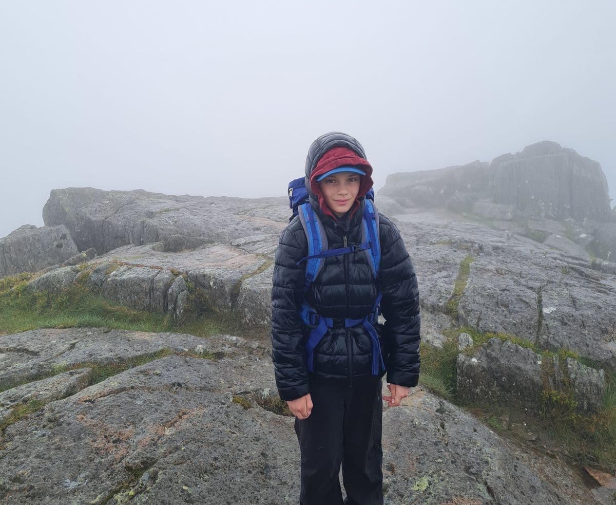 A young boy in hiking gear looks at the camera as he stands on a misty mountain top