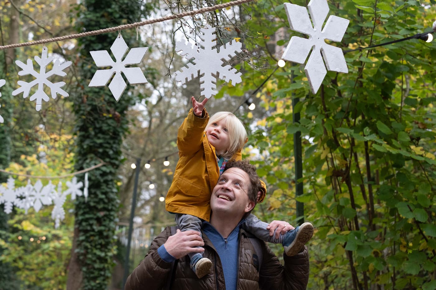 Dad with daughter on his shoulders, walking through a Festive Woodland. Daughter reaches to touch a snowflake hanging from the trees.
