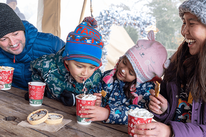 A family of four sit at a wooden bench drinking hot chocolates with marshmallows